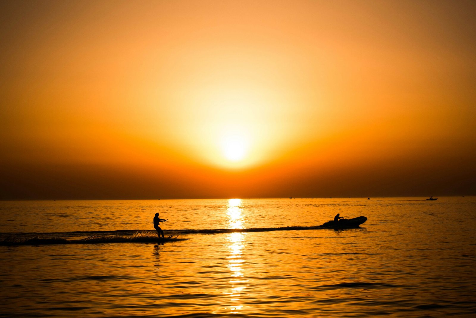 silhouette photo of man riding a motorboat with man surfboarding behind during golden hour